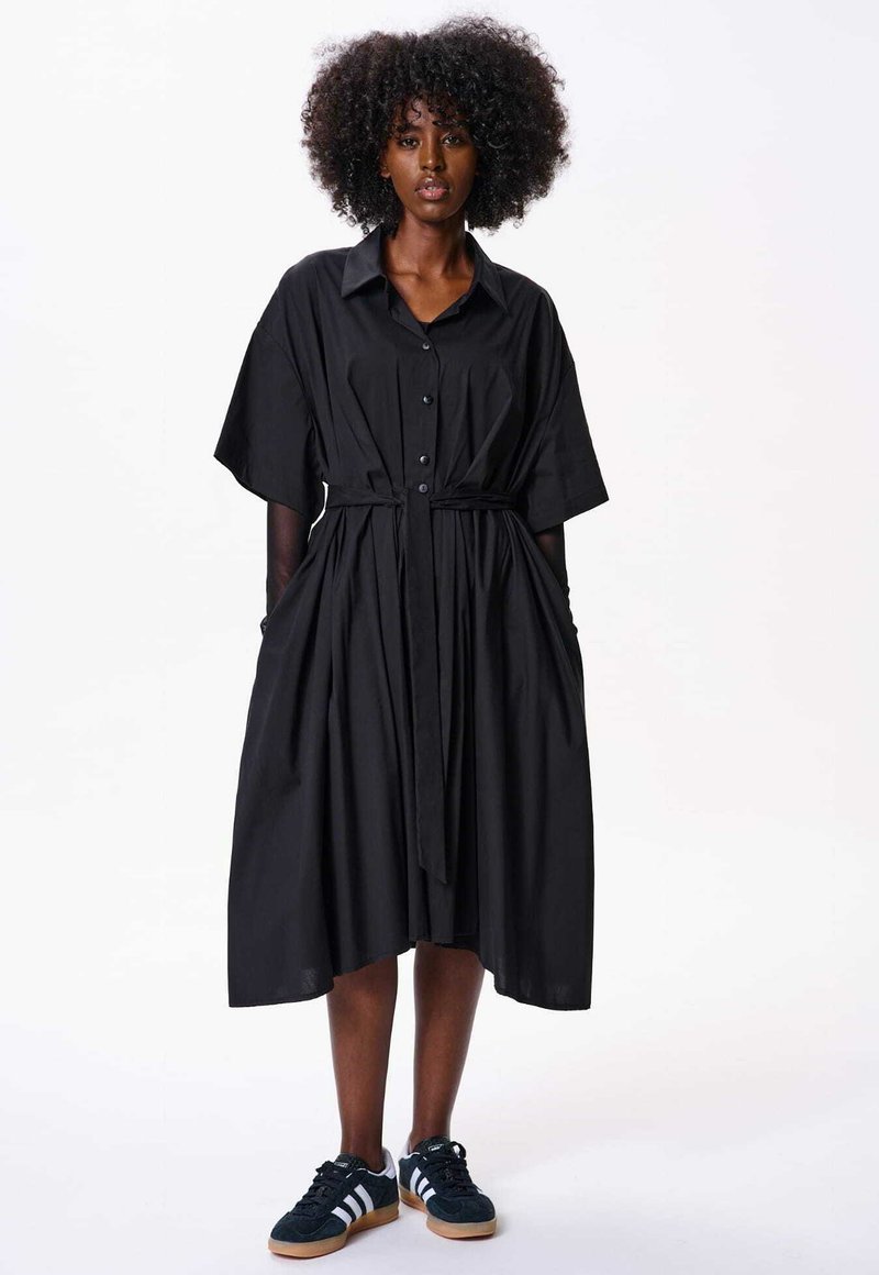 Czarna bawełniana sukienka koszulowa o oversize'owym kroju, z kołnierzykiem, zapięciem na guziki z przodu, bocznymi kieszeniami oraz materiałowym paskiem, noszona z ciemnymi trampkami.