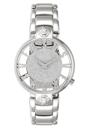 Srebrny zegarek Versus Versace z błyszczącą tarczą, otwartymi cyframi tworzącymi napis Versus, detalami głowy lwa na pasku oraz połączoną metalową bransoletą.
