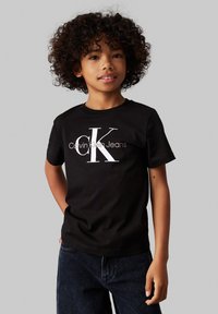 Melnas kokvilnas T-krekls ar lielu baltu "CK" logotipu un "Calvin Klein Jeans" tekstu. Vienkārša apaļa kakla izgriezne un īsas piedurknes.