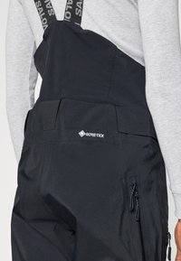 Macacão preto em Gore-Tex com alças ajustáveis, fechos laterais e uma textura suave. Apresenta um logótipo distinto nas costas.