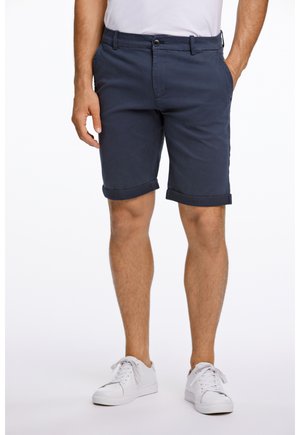 Navy blue cotton shorts with rolled hems, front pockets, and a single button closure. Paired with white sneakers and a plain white shirt.