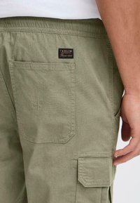 Pantalones cargo color verde oliva con una textura sutil, cinturilla elástica y una etiqueta de parche con la marca "Solid" en el bolsillo trasero.