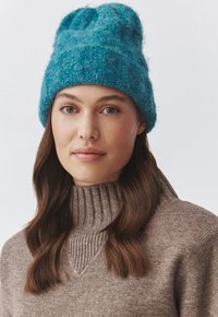 Berretto in maglia color turchese con un design a coste e finitura testurizzata. Materiale morbido e caldo e forma slouchy, indossato con un maglione color marrone chiaro.