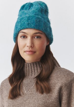 Gorro de punto en color verde azulado con un diseño acanalado y acabado texturizado. Material suave y cálido con una forma holgada, combinado con un suéter color marrón claro.