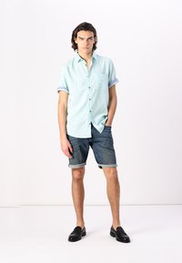 Hellblaues kurzärmliges Hemd mit einer Tasche, kombiniert mit hochgekrempelten Jeansshorts. Schwarze Mokassins runden das Outfit ab. Einfarbig weißer Hintergrund.