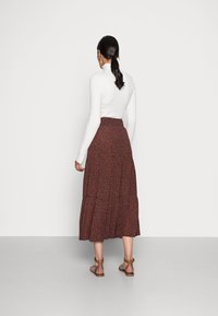 Femme avec une longue queue de cheval noire portant un haut roulé en maille blanche, une jupe midi à motifs bruns et des sandales à bride de cheville brunes, se tenant contre un mur blanc.