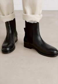 Bottines en cuir noir avec une finition lisse, dotées de panneaux latéraux élastiques et d'un petit talon arrondi. Portées avec un pantalon beige retroussé.