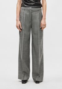 Calças xadrez cinzentas com design de perna larga e pregas, feitas de um tecido texturado, usadas com um top preto justo e sapatos pretos.