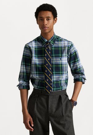 Heren geruit overhemd in groen, blauw en wit; met een button-down kraag en lange mouwen. Gecombineerd met een donkergrijze geplooide broek.