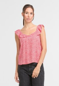 Blusa rossa con motivo floreale bianco, scollatura a V e dettagli a volant sulle maniche. Tessuto leggero, vestibilità ampia, indossata con jeans scuri.
