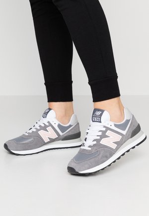 Osoba nosząca szaro-białe sneakersy New Balance 574 w połączeniu z czarnymi dopasowanymi spodniami na jednolitym jasnym tle.
