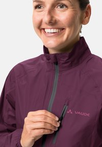 Une personne souriante en veste violette à col haut ferme la fermeture éclair de la poche poitrine de la veste.