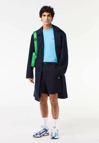 Cappotto blu navy, maglietta azzurra, pantaloncini blu navy. Borsa a tracolla verde, calze bianche e sneakers multicolori con accenti blu e rossi.