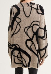 Beige ylisuurennettu neulepaita, jossa on mustia abstrakteja silmukkakuvioita. Pehmeä rakenne, lasketut olkapäät ja resorit helmassa ja hihoissa.