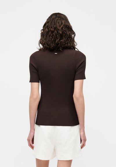 Personne aux cheveux bouclés bruns portant un haut marron foncé ajusté à manches courtes et un short blanc texturé, vue de dos sur un fond uni.