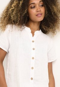 Camicia di lino bianca a maniche corte, con colletto alla cinese e bottoni in legno sulla parte anteriore. Presenta dettagli di cucitura verticale sottili.