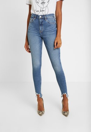 Kvinde iført lyseblå skinny jeans med højtaljede, flossede kanter og højhælede sko med slangeprint, kombineret med en hvid t-shirt med grafik.