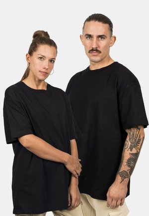 Mustat ylisuureneulokset, pyöreä kaula-aukko, lyhyet hihat, valmistettu puuvillasta. Yhdellä henkilöllä on tatuointi kädessään. Neutraali tausta.