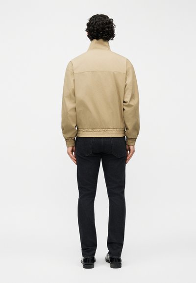 Άτομο που στέκεται με την πλάτη προς το μέρος μας, φοράει μπεζ μπουφάν, μαύρο τζιν και μαύρα παπούτσια μπροστά σε απλό ανοιχτόχρωμο φόντο.