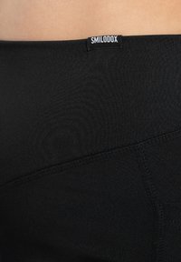 Zwarte leggings met een gladde textuur en een elastische tailleband met een wit "SMILODOX" label. Ze hebben zijnaad en een getailleerd ontwerp.