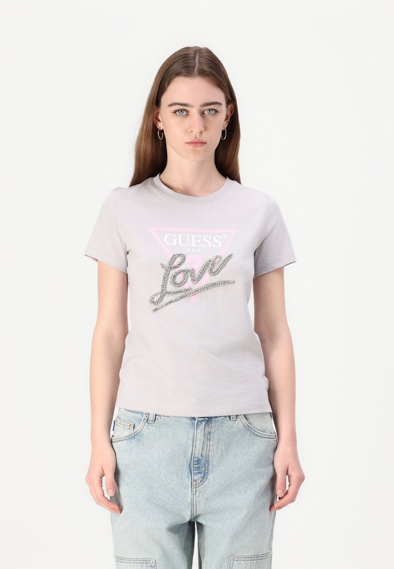 Svetlo šedé bavlnené tričko s ružovou trojuholníkovou grafikou s nápisom "GUESS" a dizajnom "Love" zdobeným kamienkami. Krátke rukávy, okrúhly výstrih.