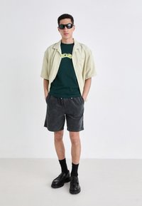 Light beige short-sleeve shirt, green graphic t-shirt, gray corduroy shorts, black shoes, and dark socks; model wearing sunglasses.