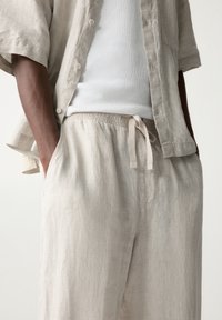 Vaaleanbeige lyhythihainen liinapaita, jossa on napit, yhdistettynä samansävyisiin väljiin housuihin, joissa on elastinen vyötärö ja nauha.