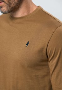 Homme portant un t-shirt marron à col rond avec une petite figure brodée en bleu marine sur le côté gauche de la poitrine, montrant les détails du cou et des épaules.