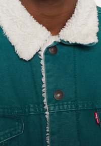 Veste Levi's teal avec col en sherpa blanc et boutons-pression marron portée par une personne à la peau foncée.