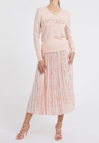 Jasnoróżowy sweter z dekoracyjnym napisem "GUESS", połączony z plisowaną spódnicą w kwiatowy wzór w delikatnych, różowych tonach i sandałkami na paski.