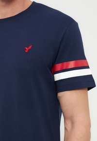 Μπλε σκούρο κοντομάνικο πουκάμισο με κόκκινες και λευκές ρίγες στο μανίκι και ένα μικρό κόκκινο λογότυπο πουλιού στο στήθος.