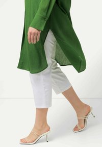 Green textured long shirt with side slits, paired with white cropped pants and beige strappy high-heeled sandals. Foot in motion.