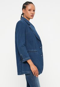 Denim blazer v temno modri barvi, s klasičnim izrezom, s pojedinačnim gumbom na zapiranju in sproščenim fitom. Ima dva sprednja žepa in rokave z gumbi.
