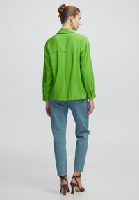 Jasnozielona koszula z długimi rękawami, kołnierzykiem i detalami na plecach, połączona z jasnoniebieskimi dżinsami oraz brązowymi sandałami na wysokim obcasie.