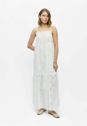 Jeune femme portant une robe maxi blanche sans manches avec des motifs floraux en broderie anglaise et des chaussures beige-rosé, debout devant un fond blanc uni.