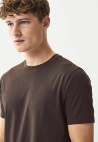 T-shirt di cotone marrone con collo rotondo, maniche corte e una texture liscia, indossato da una persona con pelle chiara e capelli ricci.