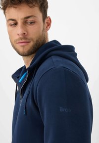 Felpa con cappuccio con zip color navy, con una texture morbida, cappuccio con coulisse e accento blu sul colletto. Presenta un logo sottile sulla manica.