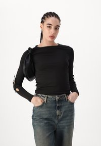 Fekete, hosszú ujjú felső, melyen redőzött anyag és kivágott részletek találhatók a könyökön, magas derekú kék farmernadrággal és fekete táskával párosítva.