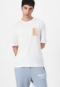 Hvid bomulds T-shirt med et guld firkantet logo mærket "HUGO" på venstre bryst. Korte ærmer og en afslappet pasform.