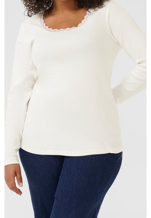 Femme portant un haut blanc à manches longues avec bordure en dentelle au niveau de l'encolure et un jean bleu foncé, debout avec une main sur la hanche.