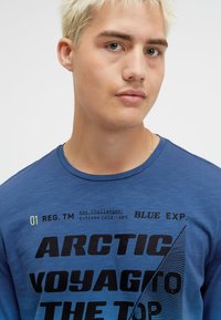 Blue crew-neck t-shirt made of soft fabric, featuring black typography with technical and geographic references, textured accents, and a slim fit.