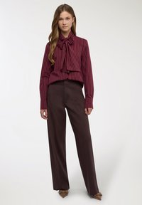 Blusa a righe color amaranto con fiocco, abbinata a pantaloni marroni larghi e tacchi appuntiti marroni. Tessuto liscio con dettagli a bottoni sulla blusa.