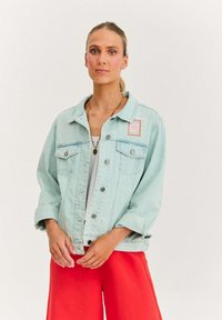 Jasnoniebieska kurtka dżinsowa z dwoma kieszeniami na piersi, srebrnymi guzikami i wyszywaną naszywką. W połączeniu z jasnoczerwonymi spodniami o luźnym kroju.