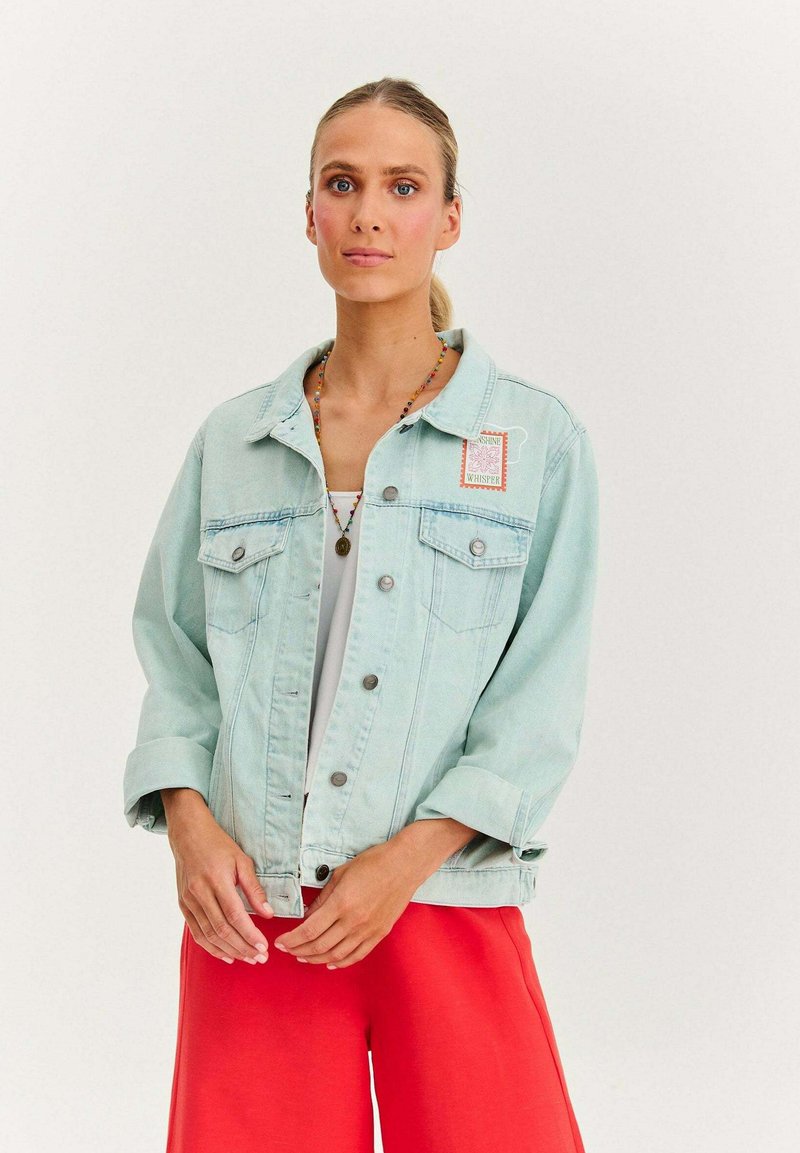 Jasnoniebieska kurtka dżinsowa z dwoma kieszeniami na piersi, srebrnymi guzikami i wyszywaną naszywką. W połączeniu z jasnoczerwonymi spodniami o luźnym kroju.