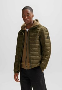Chaqueta acolchada verde oliva con cierre de cremallera, cuello alto y puños elásticos, usada sobre una sudadera marrón, combinada con pantalones negros.
