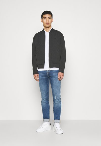 Graue Bomberjacke mit Reißverschluss, getragen über einem weißen Hemd, kombiniert mit hellblauen, ausgefransten Jeans und weißen Sneakers.
