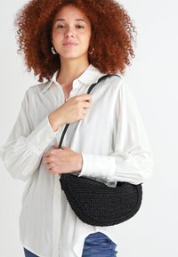 Borsa intrecciata nera con una forma curva, dotata di una sola tracolla. Il design testurizzato aggiunge dettagli, completando una camicia a strisce bianche.