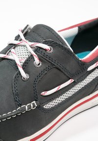 Grauer Lederschuh mit weißen Mesh-Akzenten, roten und weißen Schnürsenkeln sowie einer rot-weißen Sohle, die dekorative Nähte an den Seiten aufweist.