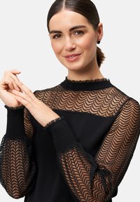 Schwarzer Pullover mit durchsichtigen, gemusterten Ärmeln und gekerbtem Rand sowie strukturierten Bündchen, der ein elegantes Design und kontrastierende Materialien hervorhebt.