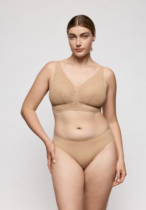 Vrouw met lichte huid en blond haar draagt een beige kanten bh en bijpassend ondergoed, staand tegen een effen lichtgrijze achtergrond.